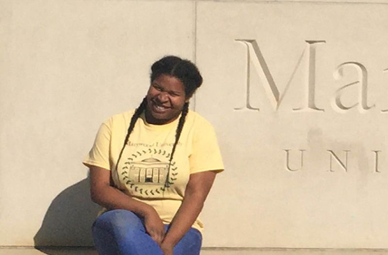 A young girl sitting in front of a stone Marywood University sign.